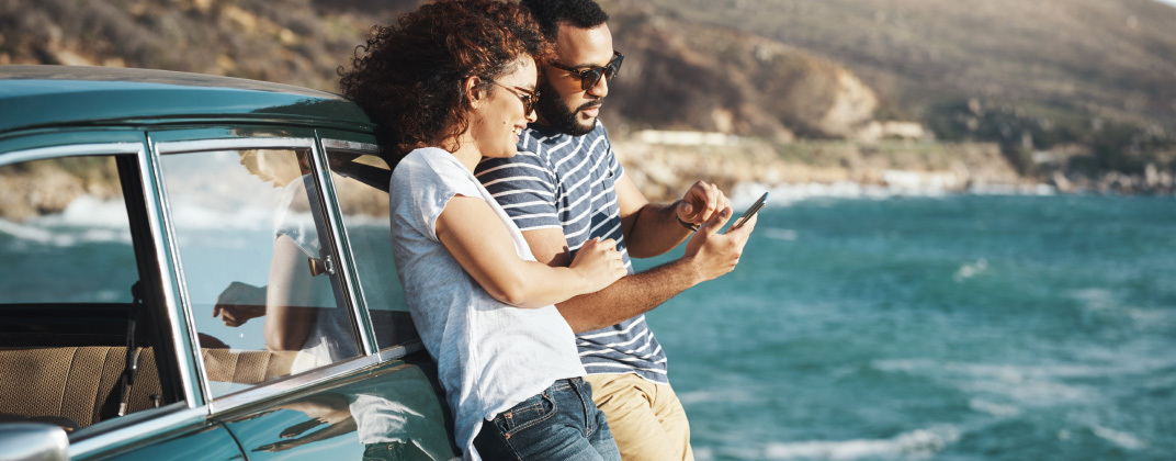 Man and woman standing next to a car next to the ocean and looking at a cell phone.